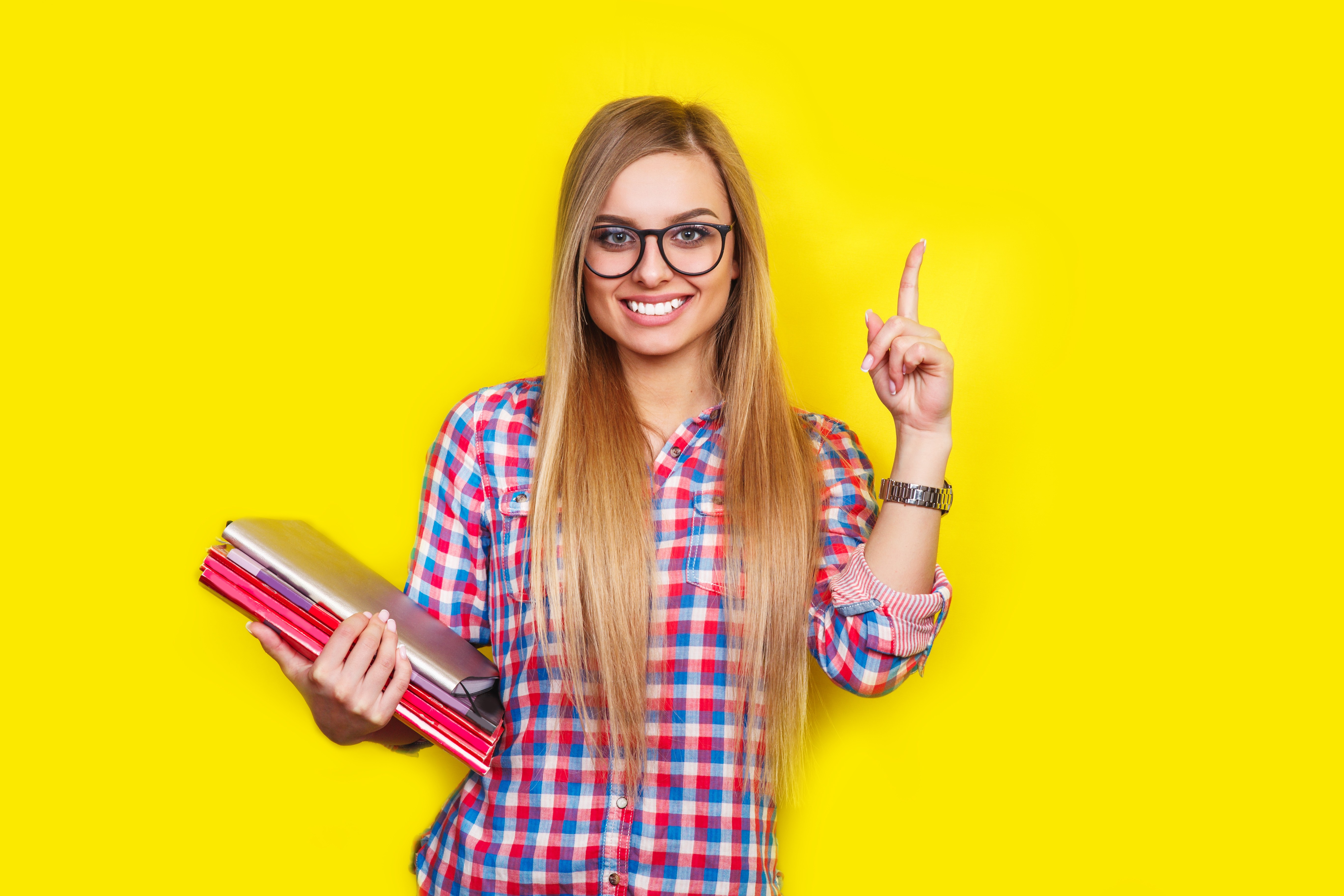 Smiling young stylish student is standing with books on yellow background in glasses and casual bright outfit
