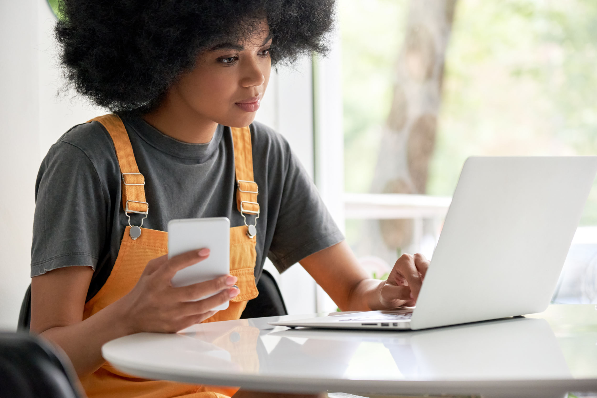 Young African woman student using laptop holding smartphone sitting at table.